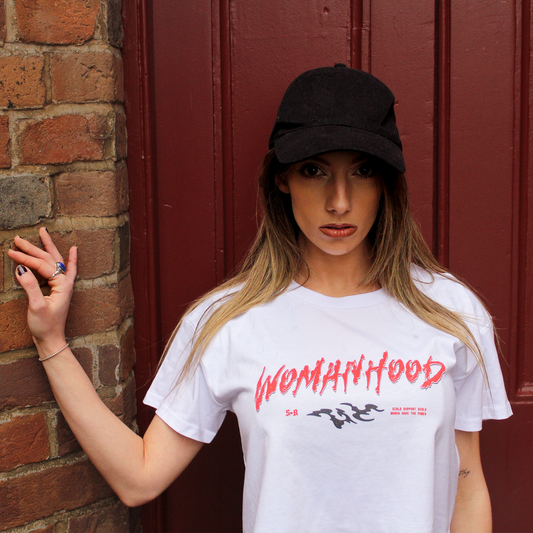 Model wearing white shirt with 'WOMANHOOD' written in bold red text. Standinging in front of a red door and brick wall wearing a black cap.